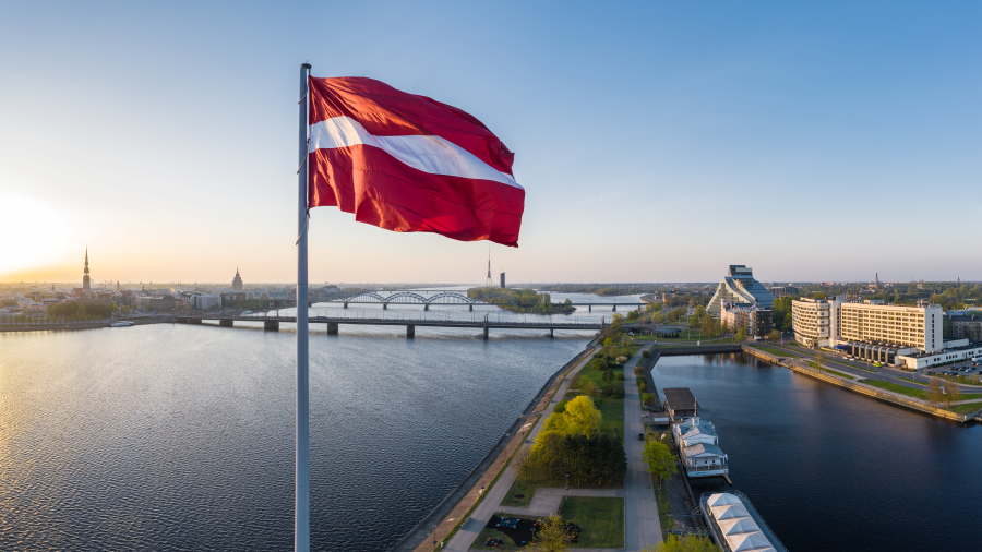 The image shows the view towards the old town of Riga, having the flag of Latvia in the foreground and the city in the background