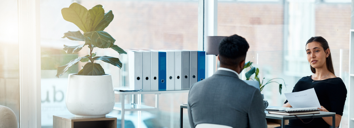 The image shows two people in an interview situation, sitting in front of each other at a desk in an office