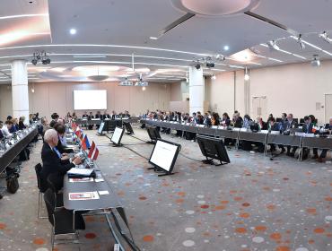 The image shows a meeting hall with multiple participants sitting at a U-shaped table group